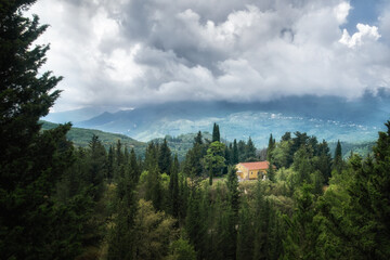 A view of a cloudy landscape on the island of Lefkada with a church in the distance surrounded by tall green trees and mountains covered with clouds.
