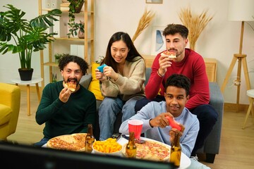 Group of multiracial friends eating pizza and playing video games in shared apartment.