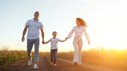 Parents lift daughter taking hands walking in field at sunset. Sun rays fall on happy family, sunlight