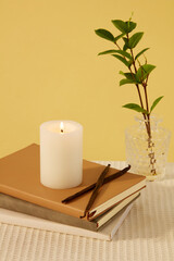 A lit candle is placed on the notebooks, next to a glass vase with green leaves on the table with yellow background. Today, most candles are made from paraffin.