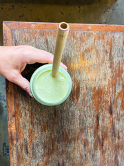 Hand holding a Green smoothie with a bamboo straw on the wooden table at the cafe