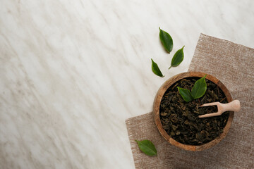 Wooden bowl with scoop and dry green tea leaves on a white marble background. Top view, copy space.