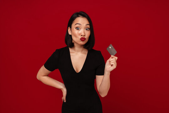 Beautiful asian woman holding credit card and making kiss lips isolated over red background