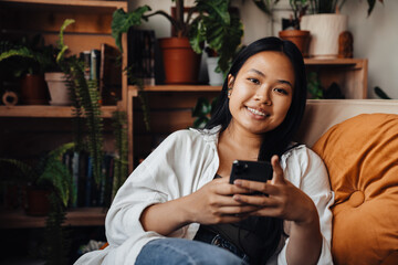 Young woman using smartphone and smiling at camera while sitting at home