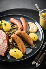 Oldenburger Grünkohl mit geräucherter Kohlwurst, Pinkel, Kassler Nacken, Kartoffeln, Senf, Petersilie auf Teller und dunkel schwarz Schiefer Hintergrund