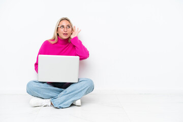 Fototapeta premium Young caucasian woman with laptop sitting on the floor listening to something by putting hand on the ear
