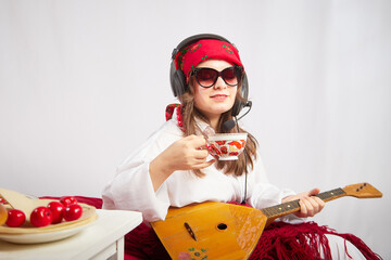 A girl in traditional national russian dress with a scarf and shawl, with a musical balalaika and modern headphones with a microphone and glasses at a set table for Easter and Maslenitsa