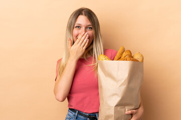 Young blonde woman holding a bag full of breads isolated on beige background with surprise facial...