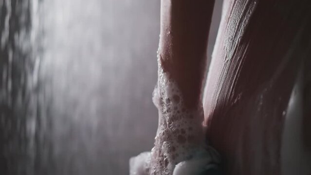 Woman washes body side with loofah in shower cabin closeup. Lady cleans body skin with foamy mesh sponge in water sprays. Pampering and hygiene