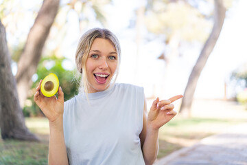 Young blonde woman holding an avocado at outdoors surprised and pointing finger to the side