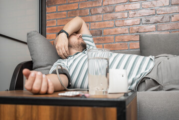 Unhealthy young man suffering from hypertension and headache, lying on the sofa at home, measures...