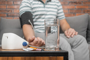 Defocused background with man sitting on sofa at home and measures blood pressure on hand with modern digital tonometer, focus on blister pack with pills on foreground