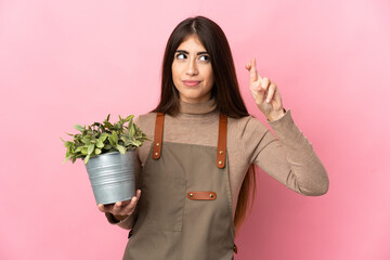 Young gardener girl holding a plant isolated on pink background with fingers crossing and wishing the best