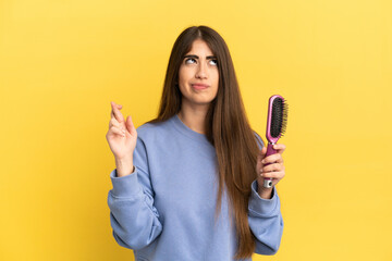 Young caucasian woman holding hairbrush isolated on blue background with fingers crossing and wishing the best