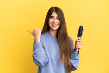 Young caucasian woman holding hairbrush isolated on blue background pointing to the side to present...