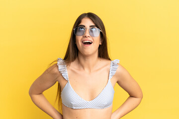 Young caucasian woman in swimsuit in summer holidays isolated on yellow background looking up and...