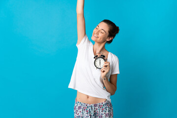Young Ireland woman isolated on blue background in pajamas and holding clock doing victory gesture