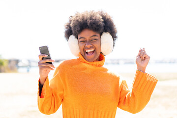 African American girl wearing winter muffs at outdoors with phone in victory position