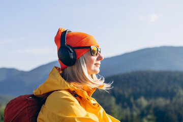 Cute smiling young woman with headphones on her head listening to the music in the mountains. Side...