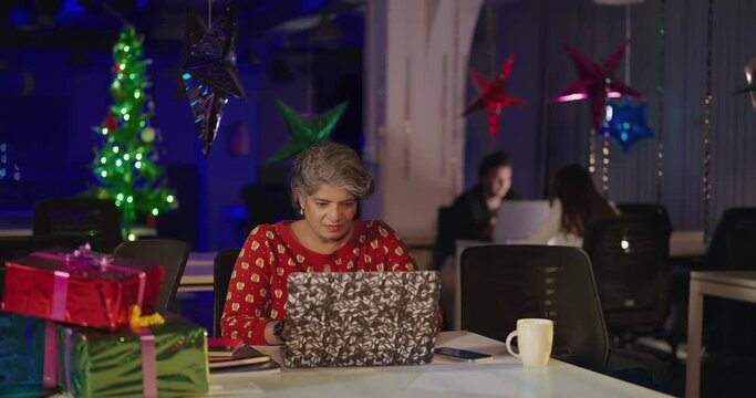 Indian Asian Hindu active mature old woman lady sitting on chair desk use laptop enjoy fun joy night new year party indoor decor office happy aged female staff do type work on computer look screen cam