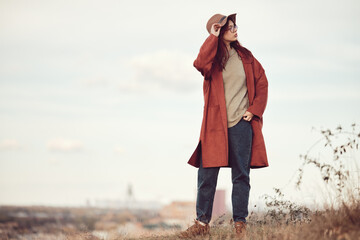 Red-haired, long-haired teenage girl stands on the grass against the backdrop of sky and cityscape, holding brim of her hat with hand. © TanyaJoy