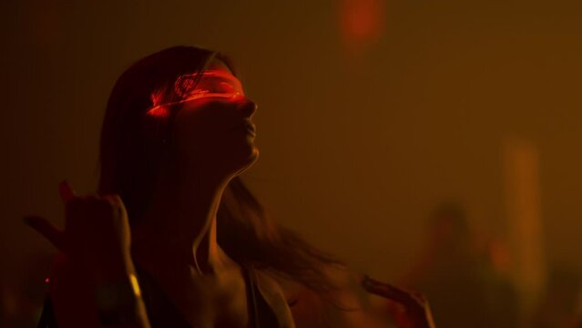 Woman in glowing glasses dancing in colorful neon light of nightclub, slow motion. Beautiful female model sensually moves to music rhytm at club party. Futuristic clubber girl on dance floor, portrait