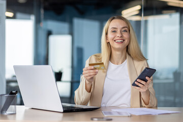 Portrait of a young businesswoman sitting in the office at a desk, smiling at the camera, holding a credit card and a phone