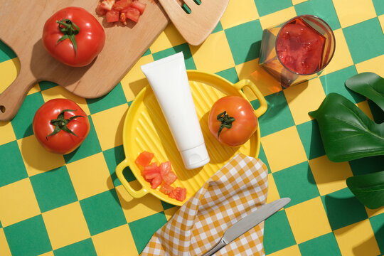 Beauty product mockup displayed on yellow ceramic pan with fresh tomatoes, glass of tomato juice, wooden tray and green leaf. Green and yellow checkered background for creative advertising