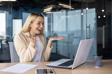 A happy young businesswoman works in the office, sits at the table and smilingly talks at an online...