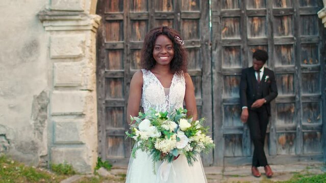 Beautiful African American fiancee with white wedding dress holding bouquet of flowers while her husband standing in background. Satisfied couple happy about creating family. Relationship concept.