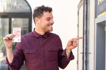 Young caucasian man taking a lot of money at outdoors pointing to the side to present a product