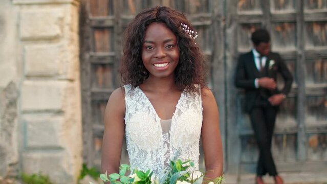 Young African American bride with amazing wedding dress holding large bouquet. Satisfied woman getting ready to starting marriage ceremony. In background attractive fiance waiting for his loving woman