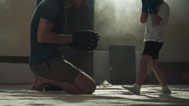 Child kicks pad on instructor hand in fighting gym. Caring trainer teaches toddler boy to hit by legs in sports center slow motion. Kickboxing lesson