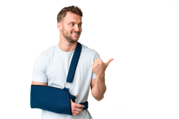 Young caucasian man with broken arm and wearing a sling over isolated chroma key background pointing to the side to present a product