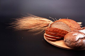 Whole and sliced breads and ears of wheat on wooden plate on black background. Photograph.