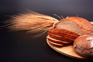 Whole and sliced breads and ears of wheat on wooden plate on black background. Photograph.