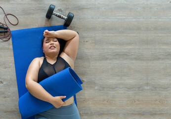 A plus-size woman lies exhausted on a blue mat, taking a breather after a vigorous workout session with dumbbell and jump rope. Top view - obrazy, fototapety, plakaty