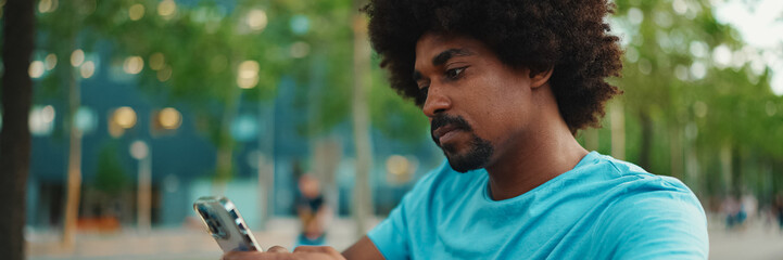 Close-up of young African American man in blue t-shirt sitting on park bench and talking on...