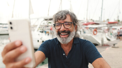 Close up, happy aged man in glasses sits in the port on yacht background uses smartphone, makes...