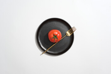 Black dish in round shaped featured a tomato and golden fork. White background. Tomato (Solanum...