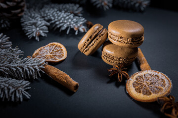Sweet chocolate macarons cookies on a dark background among dried fruits and orange slices with branches of a Christmas tree covered with snow in the background