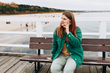 Young happy 30s woman tourist talking on smart phone and sitting on the bench outdoors. Redhead Woman walking along the waterfront, seaside. High quality photo