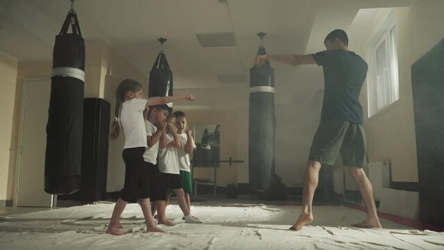 Children do punches with trainer in fighting school. Little kids learn self defence techniques with young instructor in gym slow motion. Boxing sport