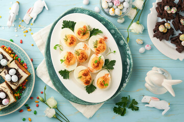 Traditional Easter brunch or dinner with stuffed eggs with paprika, carrot cake bars and chocolate nest cakes with glazed walnut eggs. Easter meal dishes with holday decorations. Top view, flat lay