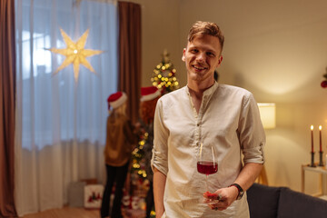Man standing with his glass and smiling while his friends decorate the Christmas tree