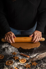 View of woman's hands with rolling pin on dark table with gingerbread cookie dough, flour, cinnamon and molds, black background, vertical