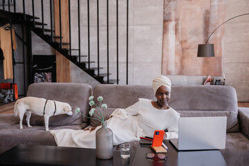 An African woman, white in traditional clothes in a turban, is lying on the sofa, working with a...