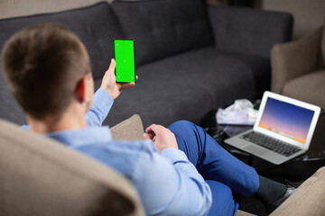 Male psychologist sitting at workplace and looking at mobile phone screen with green chromakey. Operating laptop with image on desktop - obrazy, fototapety, plakaty