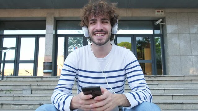 Slow Motion. Young Male University Student Sitting On Stairs Wearing Headphones, Using Phone Looking At Camera Smiling.