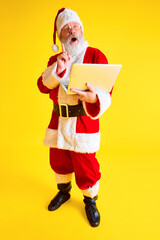 Fototapeta na wymiar Mature man with long beard looks like Santa Claus taking order online in laptop with present for children against yellow studio background.
